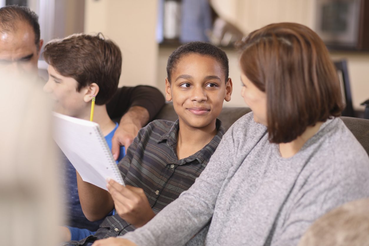 young foster child looking at foster parent while she helps him with homework on the couch